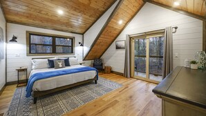 A bedroom with a sloped ceiling, a bed with a blue blanket, a window, and a view of the outdoors. - Secluded 2 Acres • 2 King Suites • Hot Tub • Sleeps 4–8 (Broken Bow)