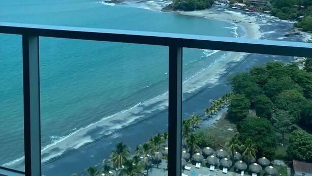 Un balcón con vista a la playa, el océano y un área de piscina con sombrillas.