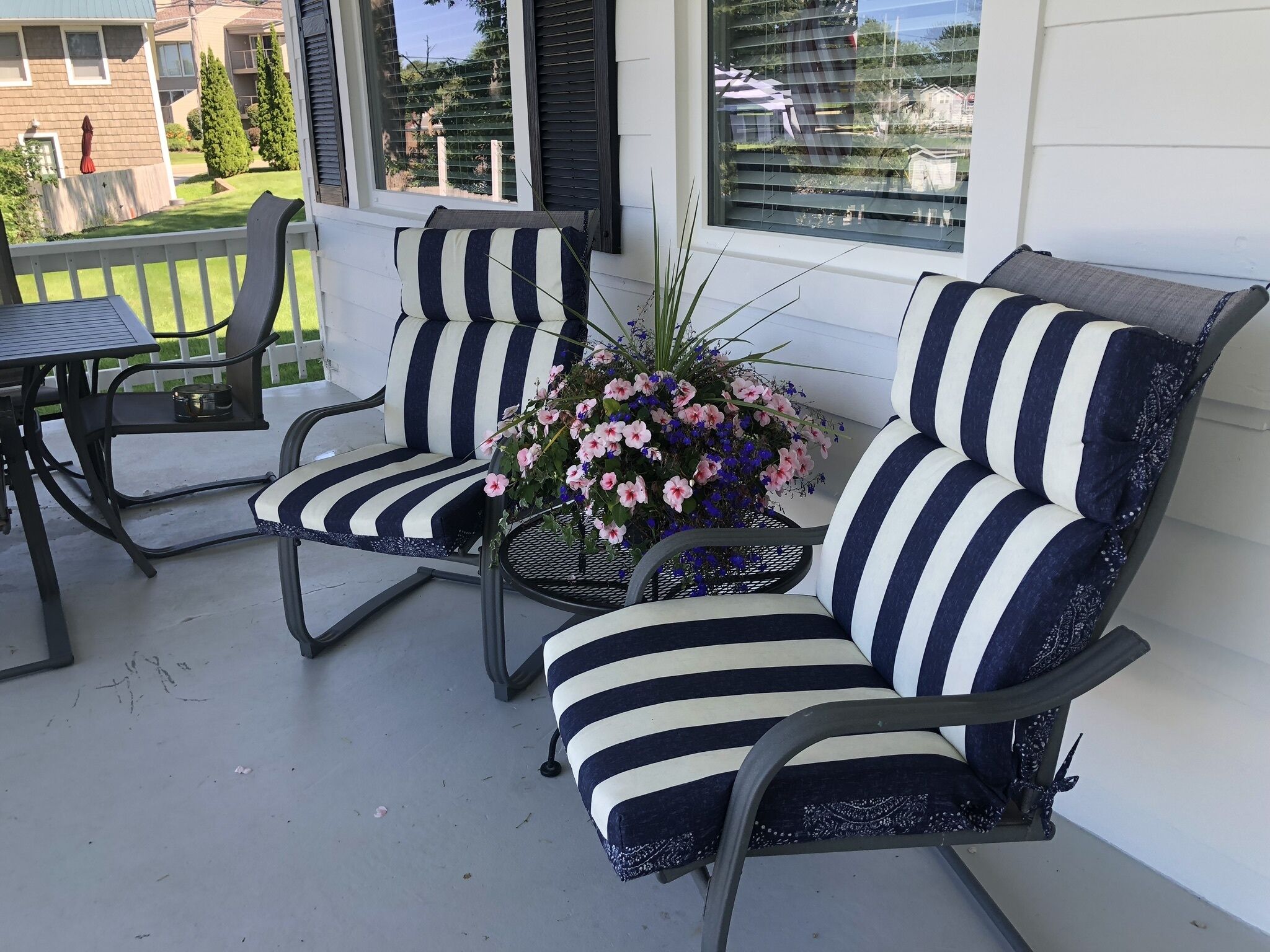 Une véranda avec deux chaises à rayures et une petite table, un pot de fleurs avec des fleurs roses, et une vue sur les maisons voisines.