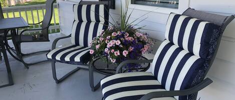 Une véranda avec deux chaises à rayures et une petite table, un pot de fleurs avec des fleurs roses, et une vue sur les maisons voisines.