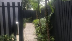 A narrow pathway leading to a building with a dark fence on one side and a palm tree on the other.