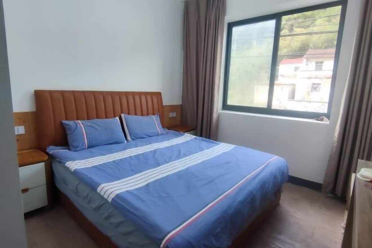 Ein Schlafzimmer mit Bett, Holz-Kopfstück, zwei Kissen, einem Nachttisch, einem Fenster mit Vorhängen und Blick auf Gebäude.