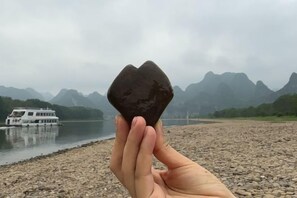 A hand holding a heart-shaped stone with a river and mountains in the background. - Fusheng Banri Guesthouse (Guilin Zhujiang) (Guilin)