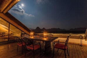 A wooden deck with a table and chairs, overlooking a beach and mountains at night. - Fusheng Banri Guesthouse (Guilin Zhujiang) (Guilin)