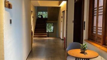 A hallway with a staircase leading to a second floor, a round table with a plant, and a window with wooden shutters.