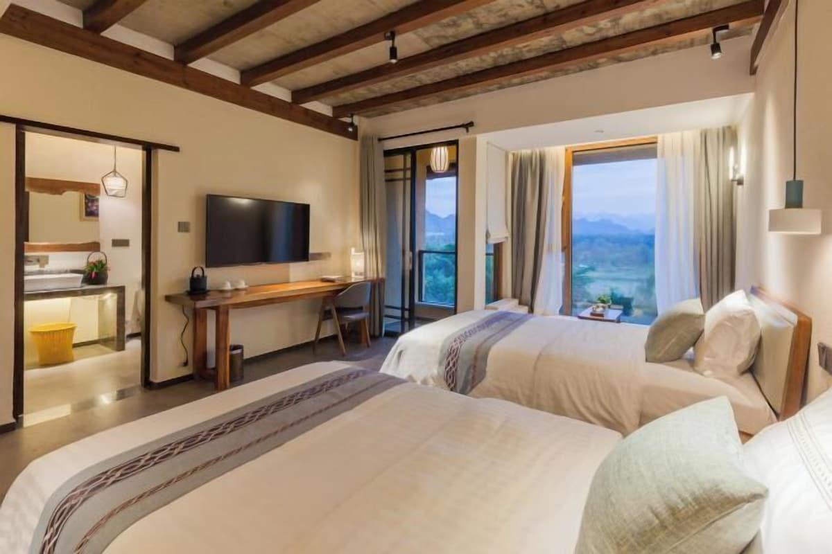 A hotel room with two beds, a desk, a TV, and a balcony with a view.