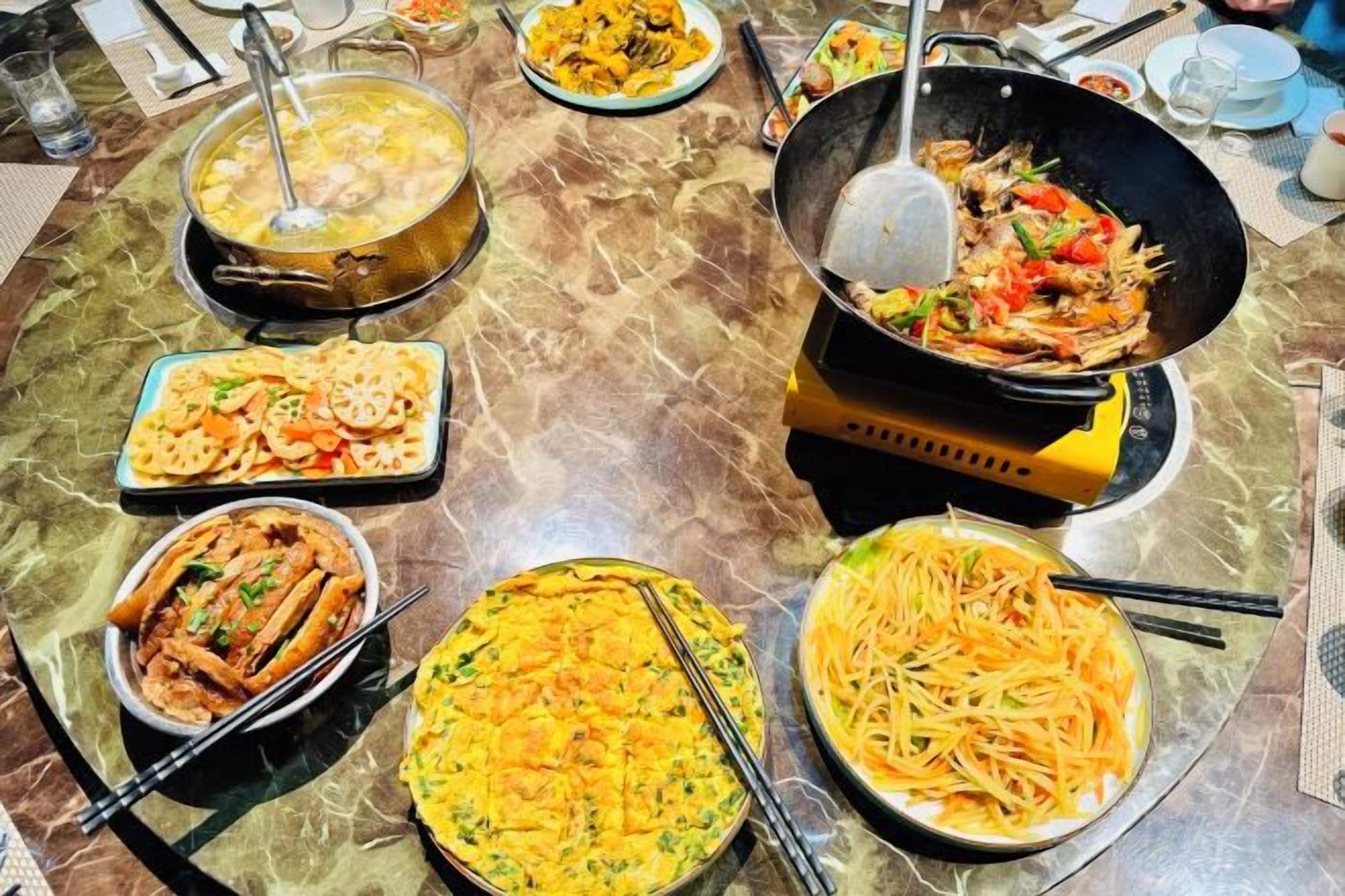 Une grande table où l’on partage un repas, avec différents plats, notamment des nouilles et des légumes sautés.