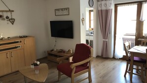 A living room with a TV, wooden furniture, a red armchair, a dining table, and a kitchen area visible through an open door.
