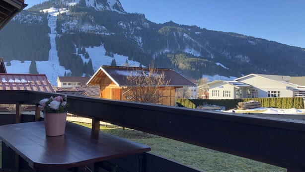A mountain view with snow-covered peaks, a wooden building, and a balcony with a table and flowers.