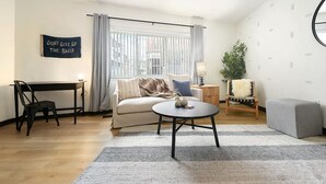 Una sala de estar moderna con un sofá, una mesa de centro redonda, un escritorio y una silla. Hay un letrero en la pared y una ventana con cortinas.