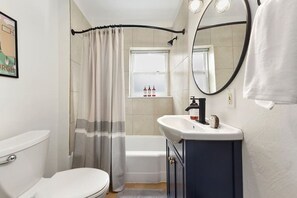A bathroom with a toilet, a sink with a mirror, a shower curtain, and a window with bottles on the sill. - Butler St Cozy Retreat, Walk to Breweries & Shops (Pittsburgh)