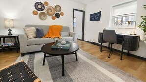 Una sala de estar moderna con un sofá gris, una mesa de centro redonda, una mesita auxiliar pequeña, un escritorio con una silla y decoración en la pared.