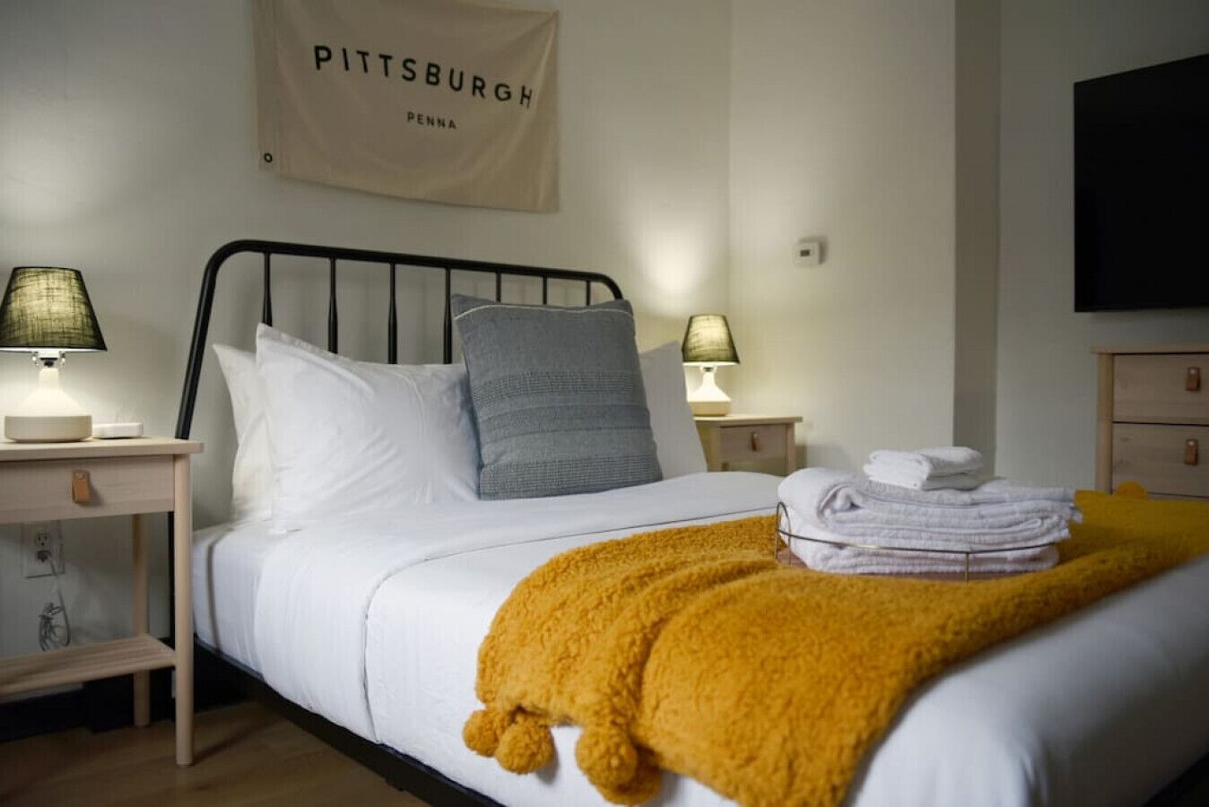 Ein ordentlich bezogenes Bett mit weißen Leinentextilien, einer senffarbenen Tagesdecke und einem Kopfkissen. Zwei Nachttischlampen, ein an der Wand befestigtes Schild mit der Aufschrift "Pittsburgh Penna" sowie ein Fernseher an der Wand sind ebenfalls zu sehen.