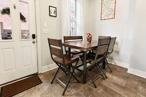 Un coin repas avec une table en bois et quatre chaises, une porte blanche avec un paillasson, et une fenêtre avec des rideaux.