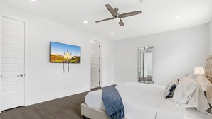 A bedroom with a large bed, a ceiling fan, a wall-mounted TV, and a door leading to another room. - New Constructed Luxury 3-BD House in Mid-City (New Orleans)