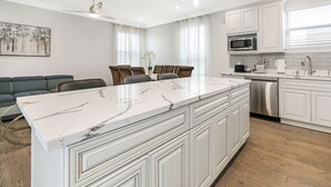 A modern kitchen with white cabinetry, a marble island, and a dining area with a gray sofa and chairs. - New Constructed Luxury 3-BD House in Mid-City (New Orleans)