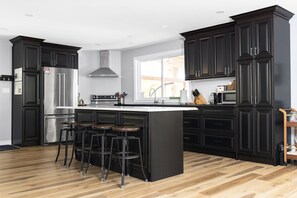 A modern kitchen with dark cabinetry, a stainless steel refrigerator, a large island with stools, and a window providing natural light. - Modern cottage escape in Muskoka (Huntsville)