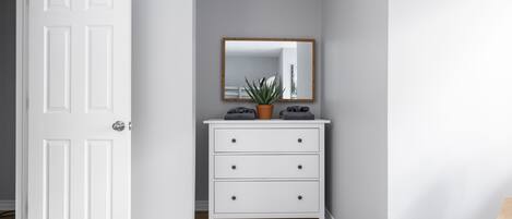 Un pasillo con una puerta blanca, un aparador blanco con cajones, un espejo y una planta en maceta.