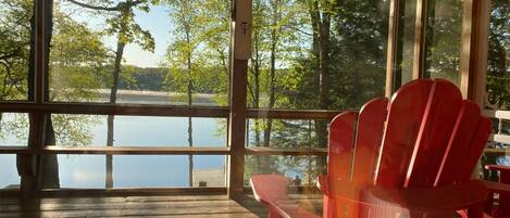 Une terrasse en bois, agrémentée d’une véranda vitrée donnant sur un lac, avec des chaises Adirondack rouges et une table.