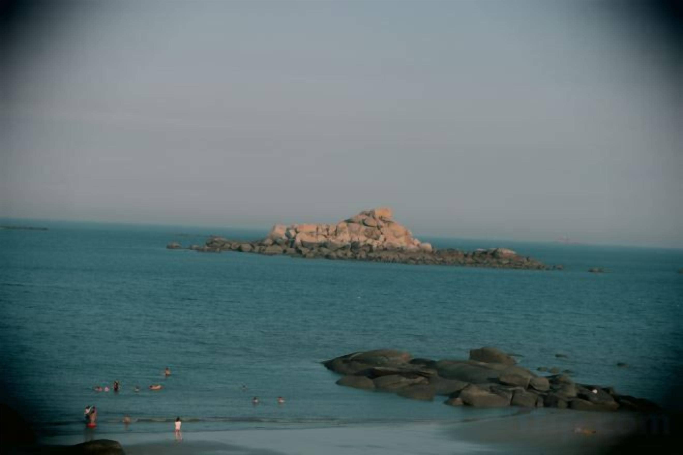 A coastal scene with a rocky outcrop in the water and a beach with people.