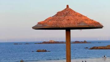 A beach umbrella with a thatched roof, a wooden post, and a view of the sea with rocks and wind turbines in the distance.