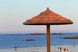 A beach umbrella with a thatched roof, a wooden post, and a view of the sea with rocks and wind turbines in the distance.