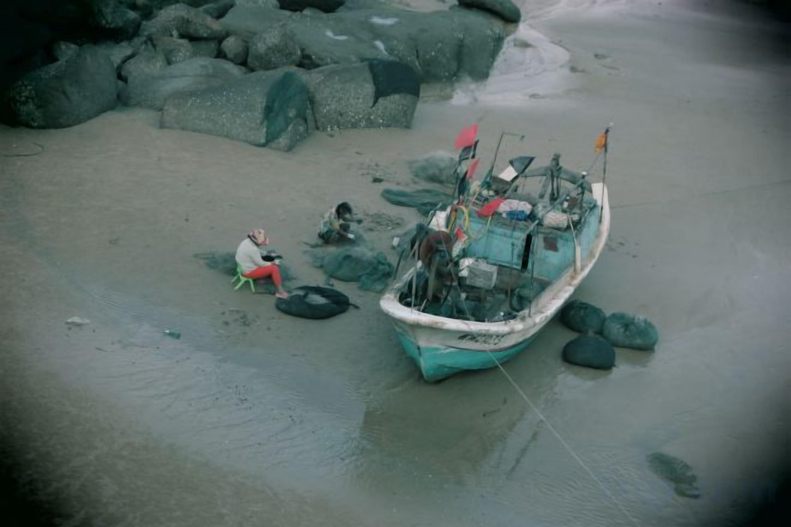 En båt på en sandstrand med klippor, en person som sitter på marken och en flagga på båten.