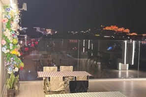 A rooftop terrace with tables and chairs, a decorative floral arrangement, and a city view at night.