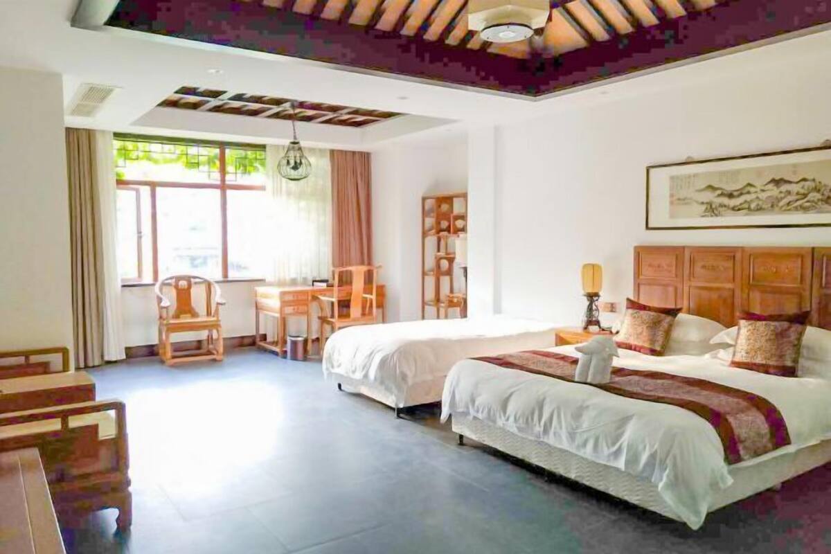 Ein Hotelzimmer mit zwei Betten, einem Schreibtisch und einem Stuhl. Es gibt ein Fenster mit Vorhängen, eine Decke mit sichtbaren Holzbalken und ein Bild an der Wand.