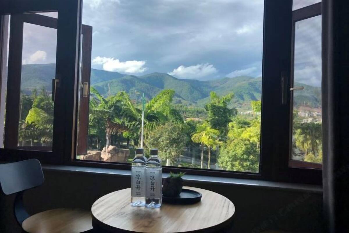 A room with a view of mountains and greenery, a round wooden table with two bottles and a small plant.