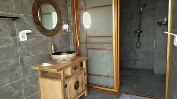 Um banheiro com toucador de madeira, espelho redondo, pia, box de vidro para chuveiro e piso revestido de azulejos.