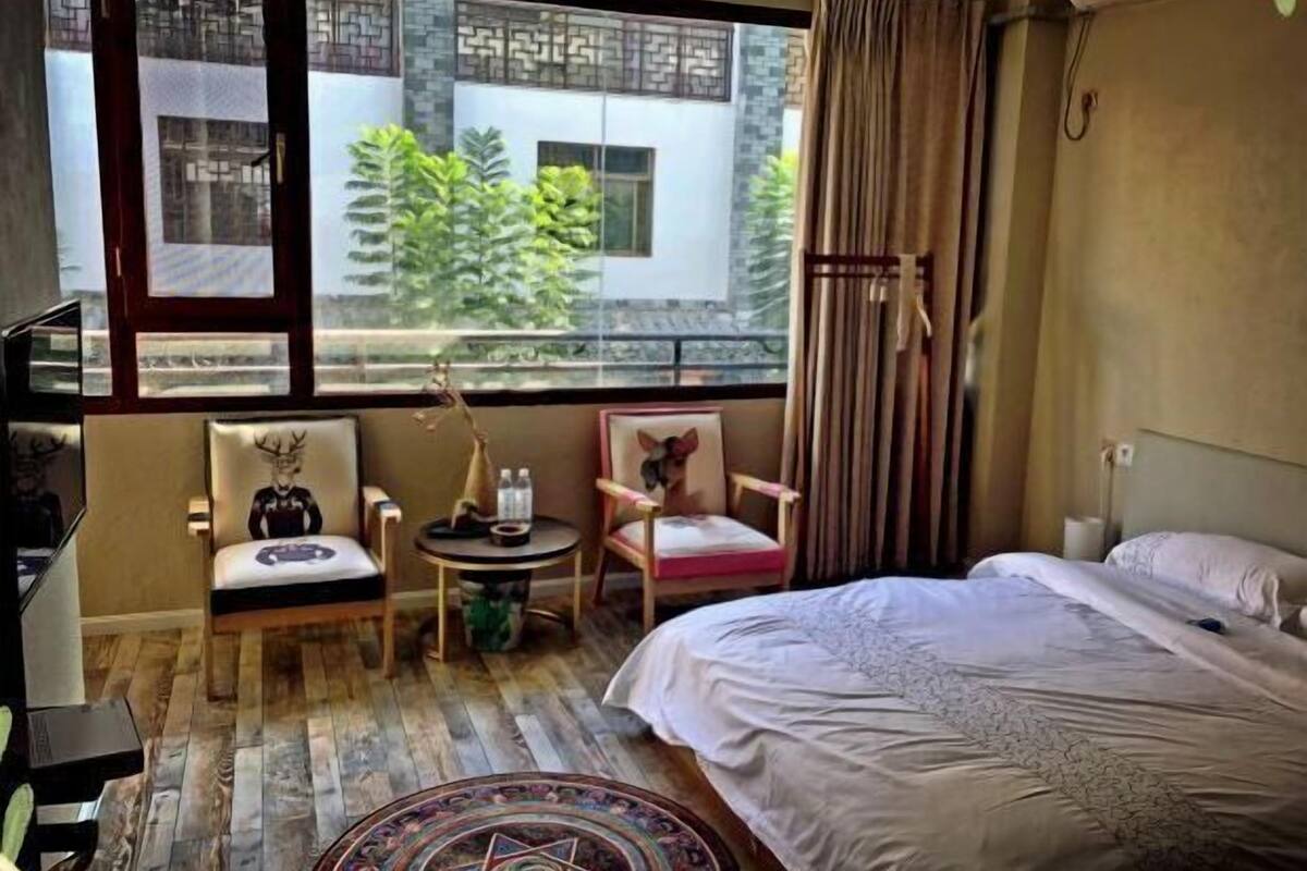 A hotel room with a bed, a chair, a small table, and a window with a view of greenery.