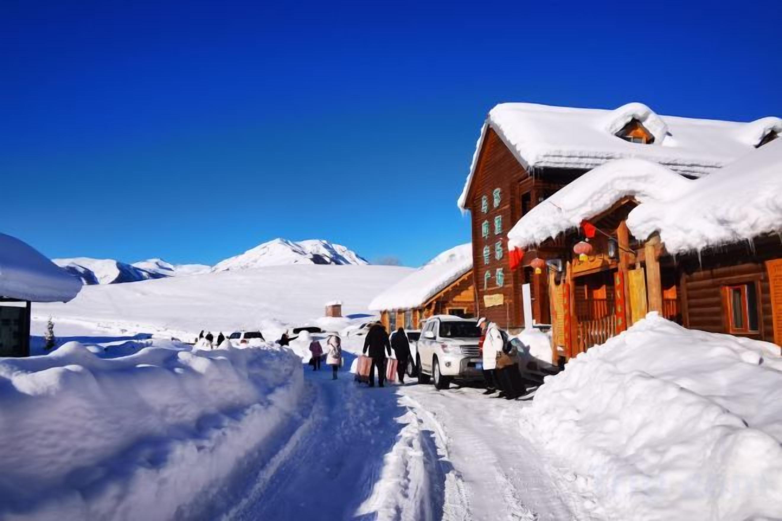 A snow-covered wooden building with a clear sky, a snow-covered road, and a mountainous background.