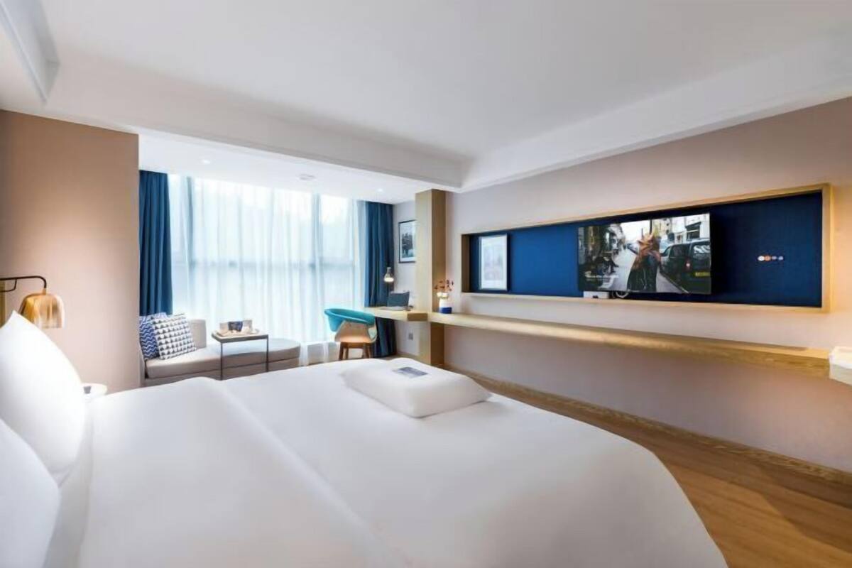 A modern hotel room with a large bed, a TV mounted on the wall, a small desk, and a window with curtains.