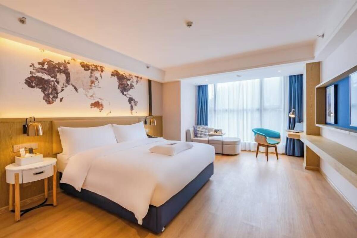 A modern hotel room with a large bed, a desk, a chair, and a world map on the wall.