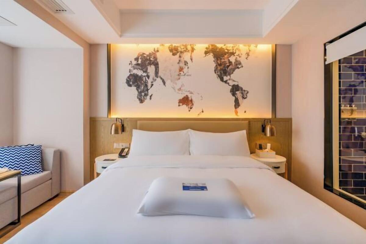 A hotel room with a large bed, a world map artwork on the wall, a bedside table with a lamp, and a view of the city through a window.