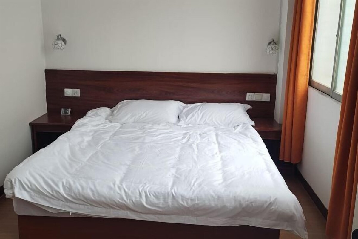 A neatly made bed in a room with a wooden headboard, a nightstand with a lamp, and a window with curtains.