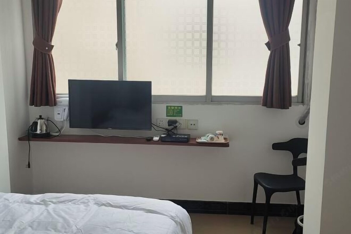 A hotel room with a bed, a TV on a stand, a desk with a computer, a chair, and a window with curtains.