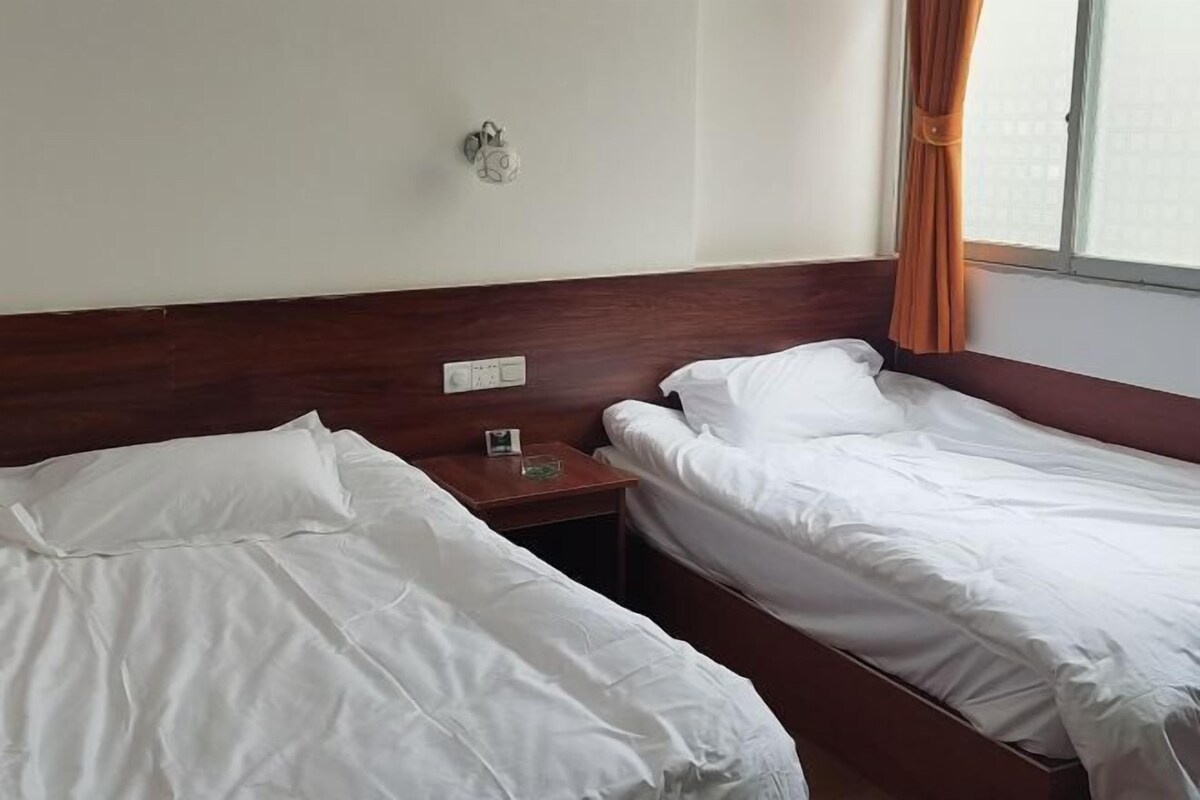 A hotel room with two single beds, a nightstand, a wall clock, and a window with curtains.