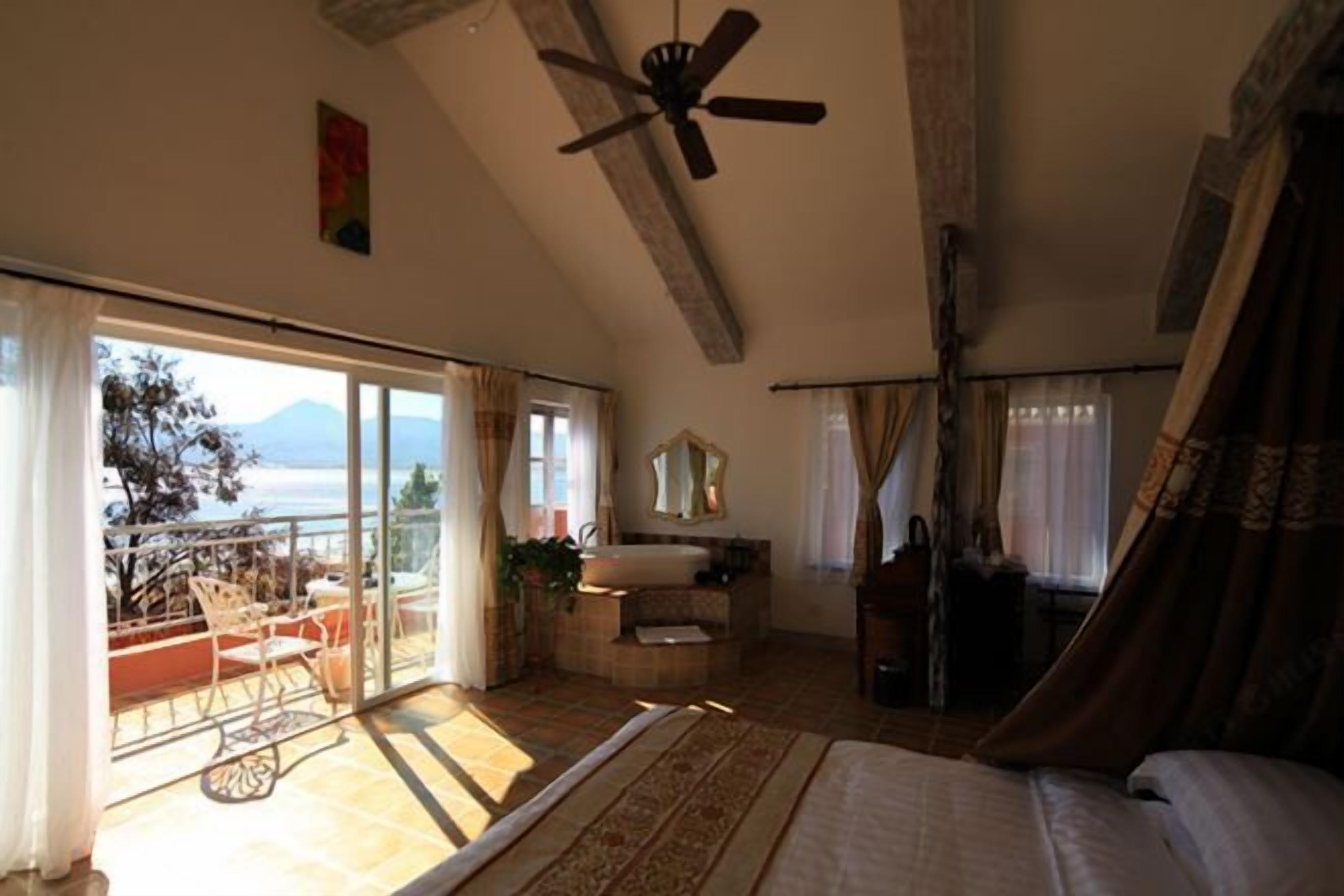 A bedroom with a large bed, a ceiling fan, a bathroom with a bathtub, and a balcony with a view.