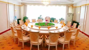 A round dining table with chairs, surrounded by a patterned carpet, with a chandelier above and a window with curtains in the background.