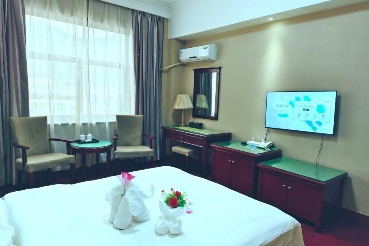 A hotel room with a bed, two chairs, a desk, a TV, and an air conditioning unit.