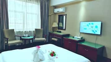 Quarto de hotel com uma cama, duas cadeiras, uma escrivaninha, televisão e ar condicionado.