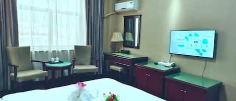 A hotel room with a bed, two chairs, a desk, a TV, and an air conditioning unit.