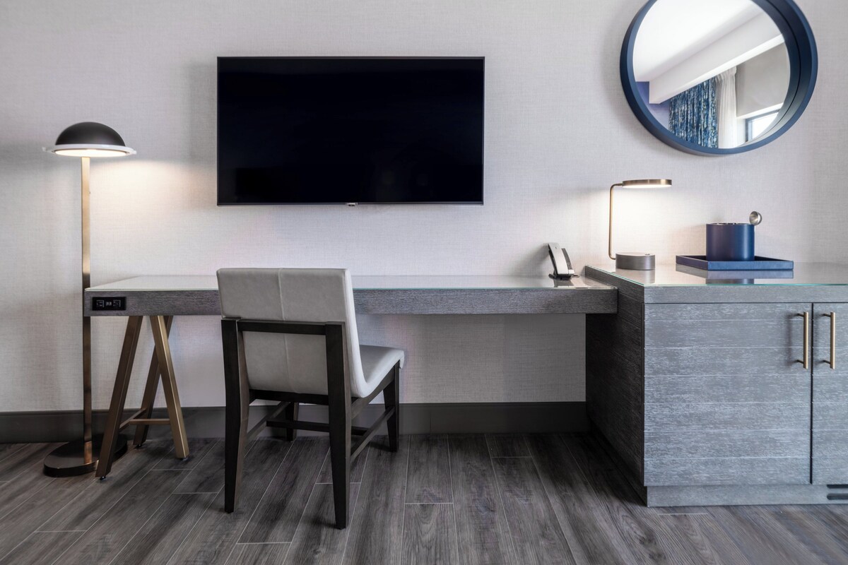 A modern hotel room with a flat-screen TV mounted on the wall, a desk with a chair, a round mirror, and a cabinet with drawers.