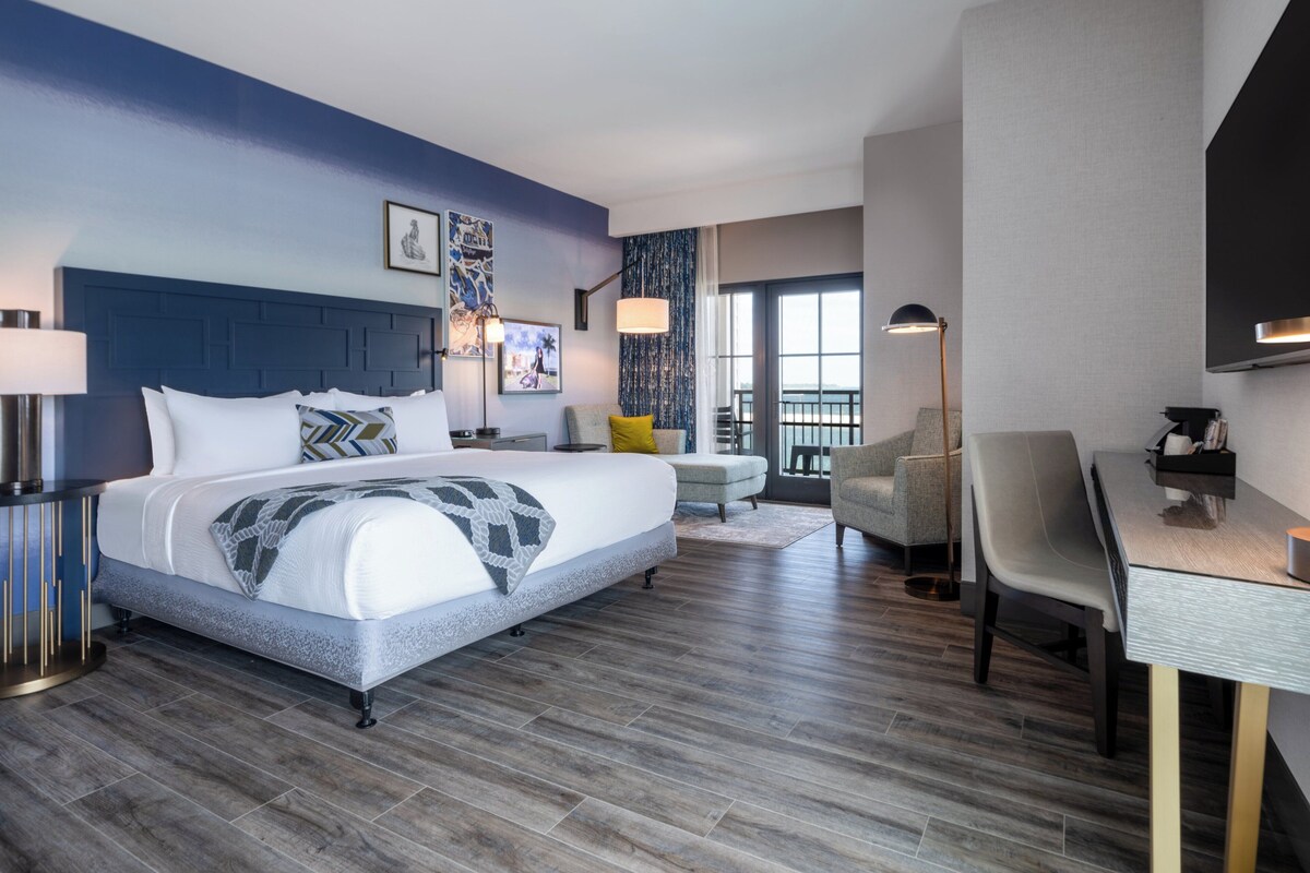 A modern hotel room with a large bed, a desk, a chair, and a balcony with a view.