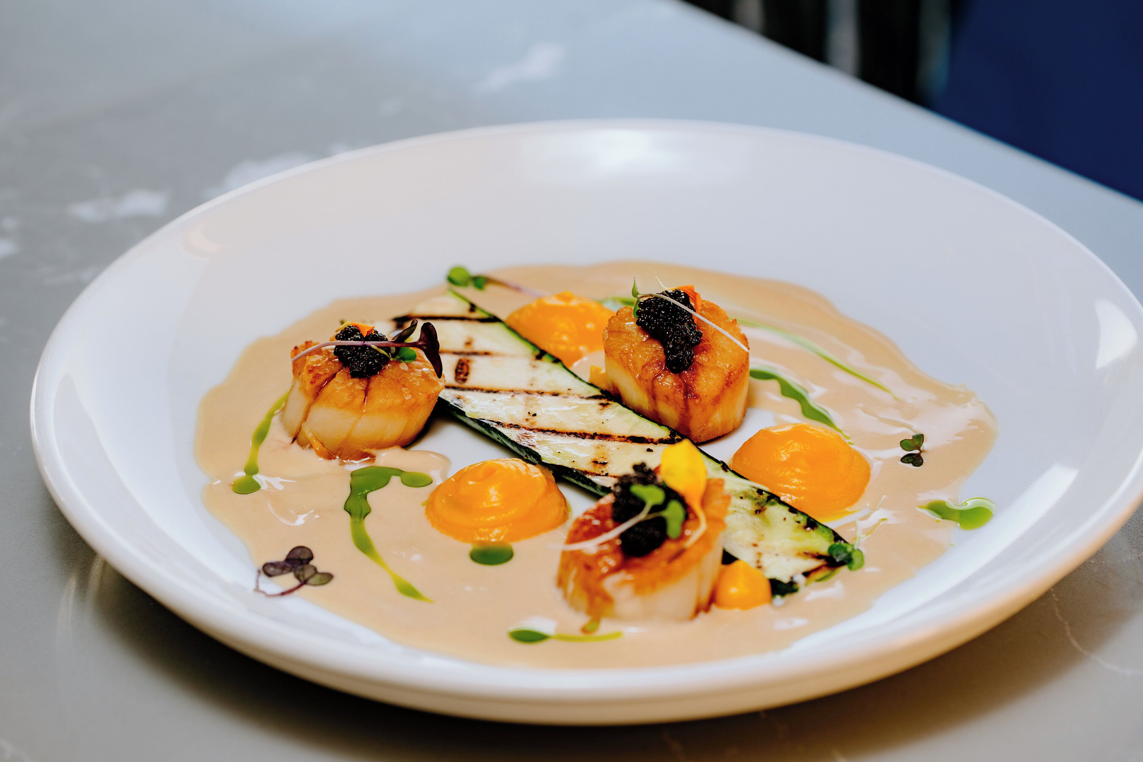 A white plate with a seafood dish, including grilled fish and vegetables, served with a creamy sauce.