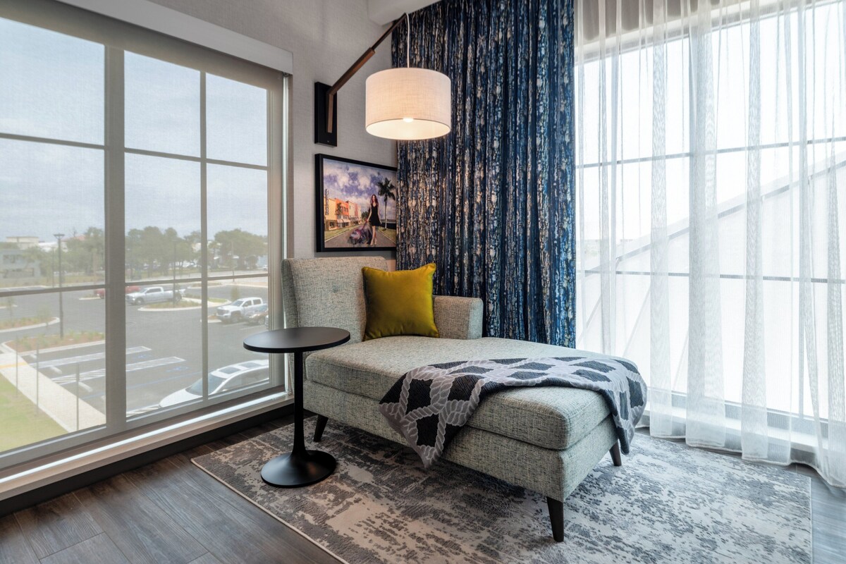 A hotel room with a large window, a sofa with a throw blanket, a small round table, and a lamp.