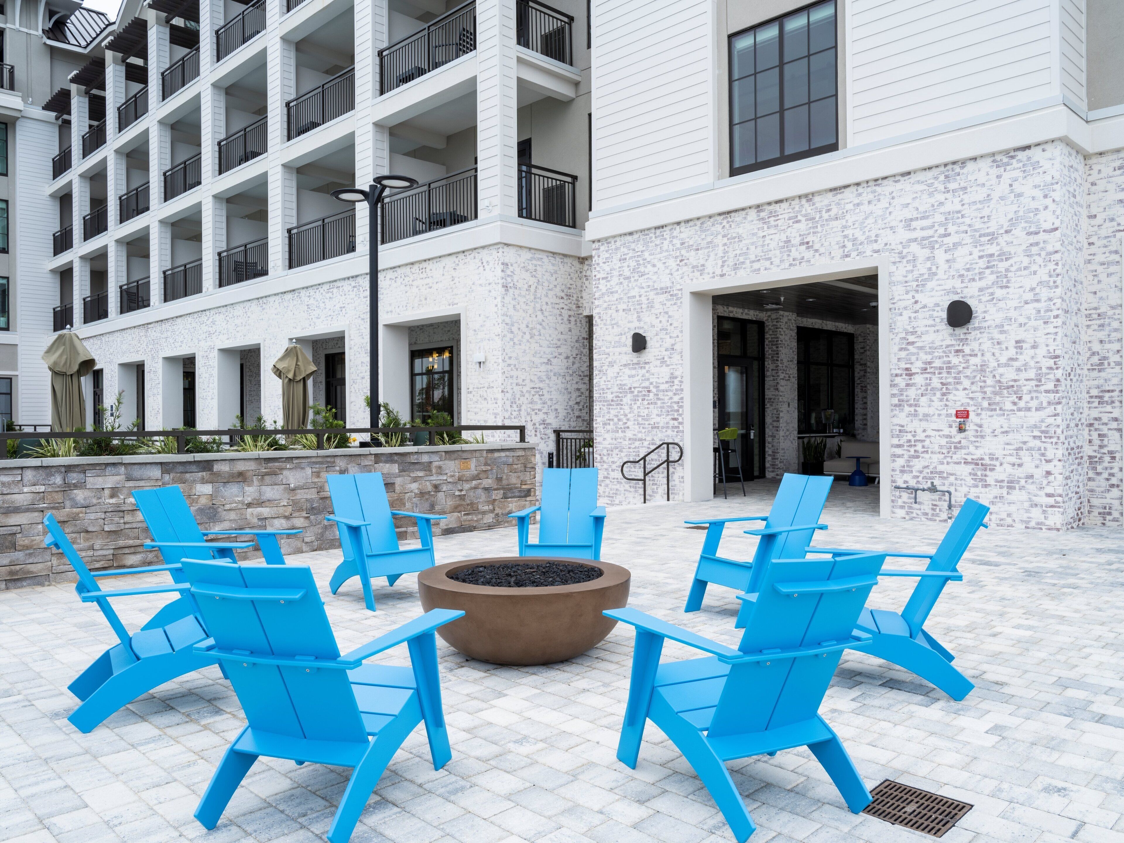 A courtyard with blue chairs arranged around a fire pit, a stone wall, and a building with multiple balconies and windows.