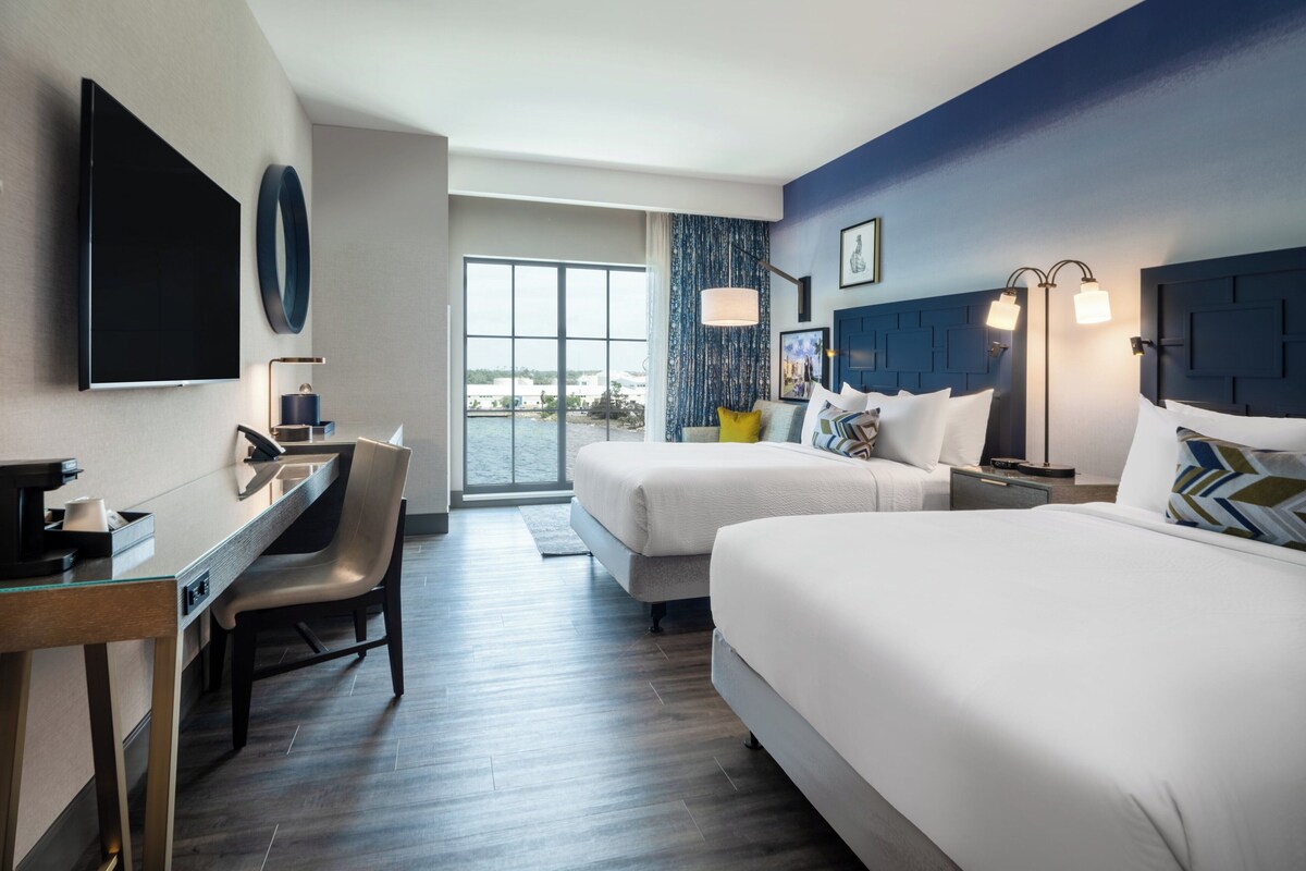 A hotel room with two beds, a desk, a chair, a TV, and a large window.
